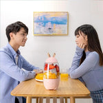 Two people sitting at a table with a pink fruit juicer between them, smiling.