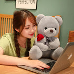 Woman using a laptop with a large teddy bear holding a rose on her lap.