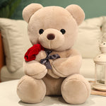 Beige teddy bear holding a red rose with a bow on a soft surface.